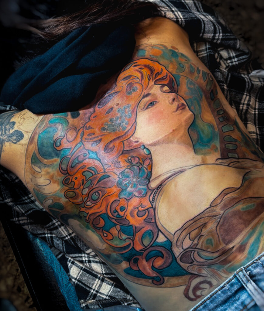 Tatouage féminin art nouveau en couleur sur le dos, lignes décoratives et palette chaude, inspiration Alphonse Mucha, réalisé par Tamara Chaudesaigues.