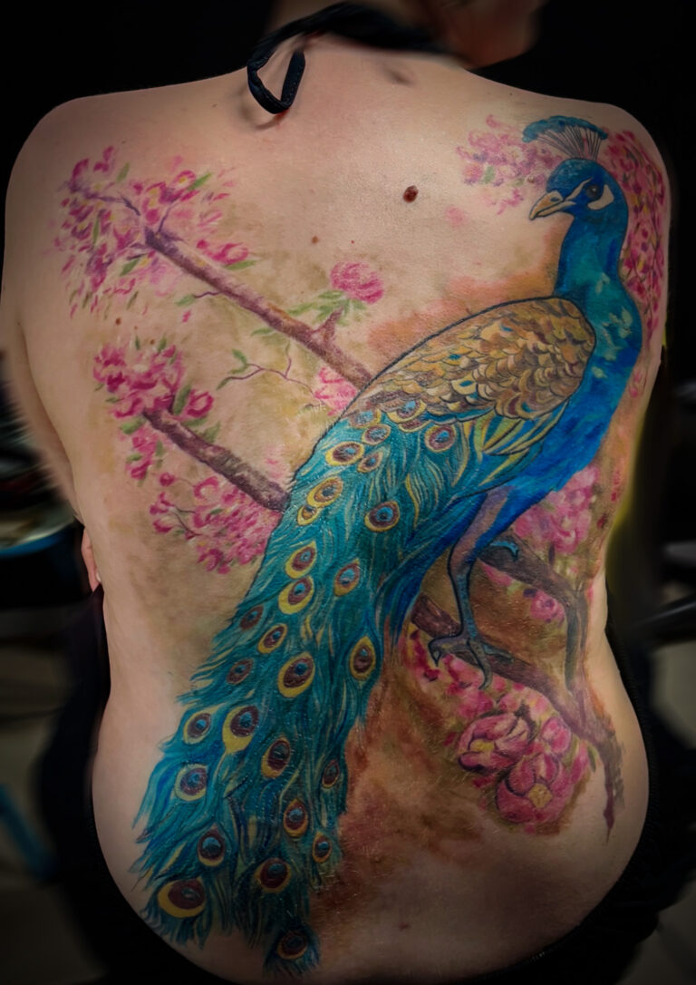 Tatouage de paon en couleur dans le dos, composition florale et branches en fleurs, inspiration art nouveau, réalisé par Tamara Chaudesaigues.