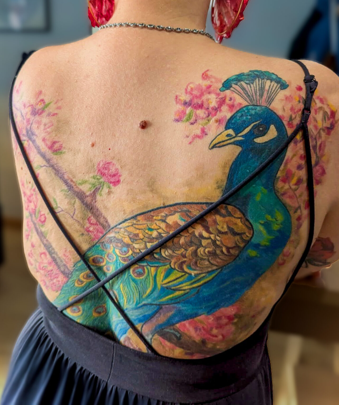 Tatouage de paon en couleur sur le dos, composition florale et lignes fluides, réalisé par Tamara Chaudesaigues.