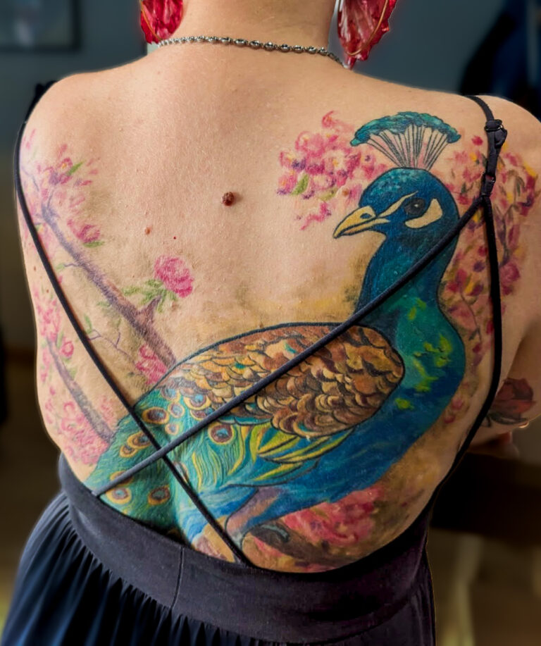 Tatouage de paon en couleur sur le dos, composition florale et lignes fluides, réalisé par Tamara Chaudesaigues.