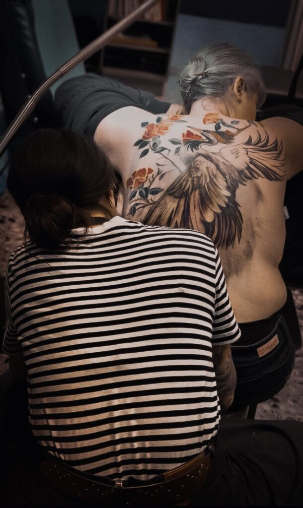 Tamara Chaudesaigues tatoueuse en séance sur un tatouage dos couleur grue et fleurs, Graphicaderme Avignon